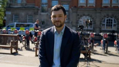 BBC's Henry Zeffman poses for the camera from a street in London on a sunny day. He wears a blazer and a light blue shirt with the top button undone.