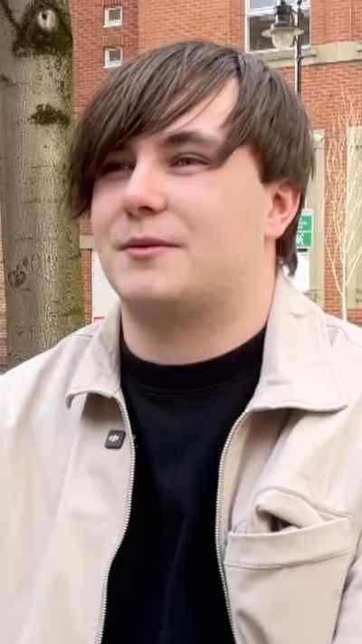 Young man with long brown hair wearing a light jacket and black t-shirt underneath
