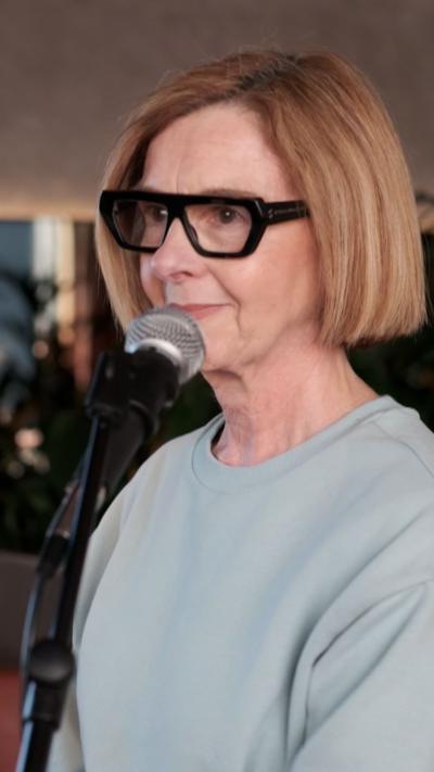 A woman with a blonde bob, black framed glasses and light green jumper is standing in front of a mic.