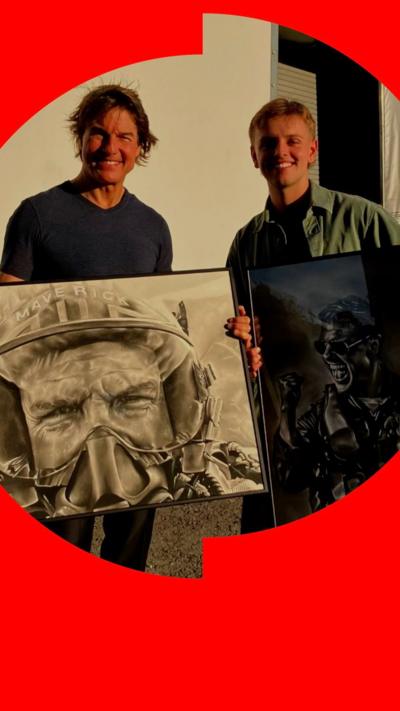Actor Tom Cruise standing next to artist Wilf Elliott. They are outside and standing in front of a white wall, both holding framed charcoal portraits of Cruise.