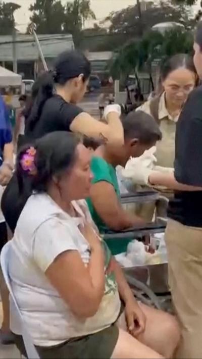 A woman is seated holding her hand to her chest, while a man seated next to her has his head looked at by a nurse wearing a back shirt. 