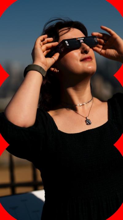 A woman wears eclipse sunglasses as she smiles and looks up at the sky