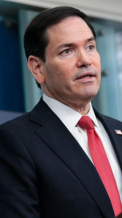 Marco Rubio wears a navy suit with a red tie and an American flag pin as he speaks to the press (who are off camera) in the White House briefing room.