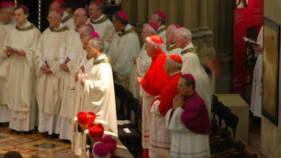 The future pope is in the foreground of a line of priests in their vestments in front of two two cardinals in red skull caps. The sun is shining on him.