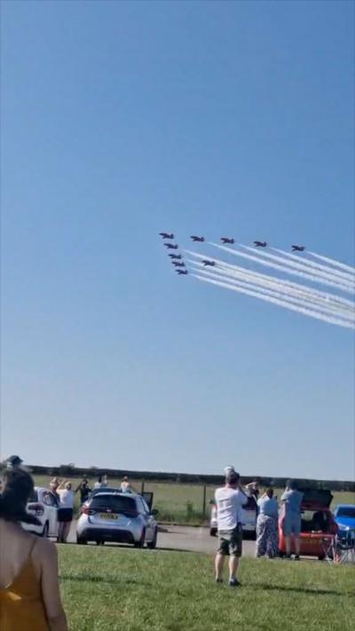 Red Arrows aircraft flies in a ten formation with white smoke trail out the back, blue sky and crowds looking from the ground.