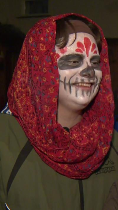A man and woman, both wearing green coats with halloween face make upo
