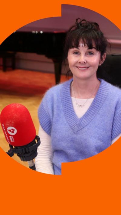 A woman smiles while sitting behind a micrpphone. She wears a light purple vest and has a brown fringe.