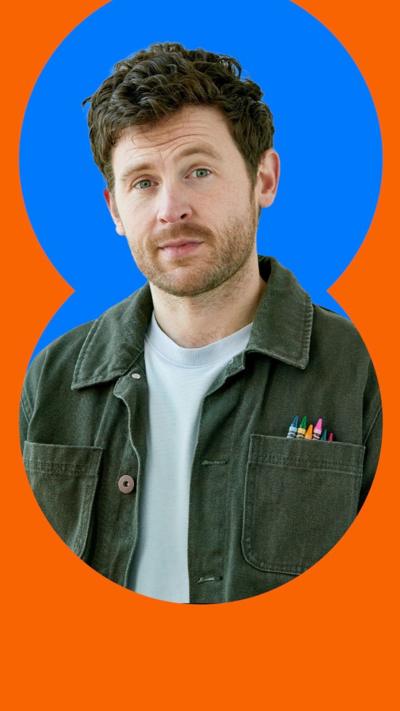 George Lewis has short brown hair and stubble and stands against a blue background