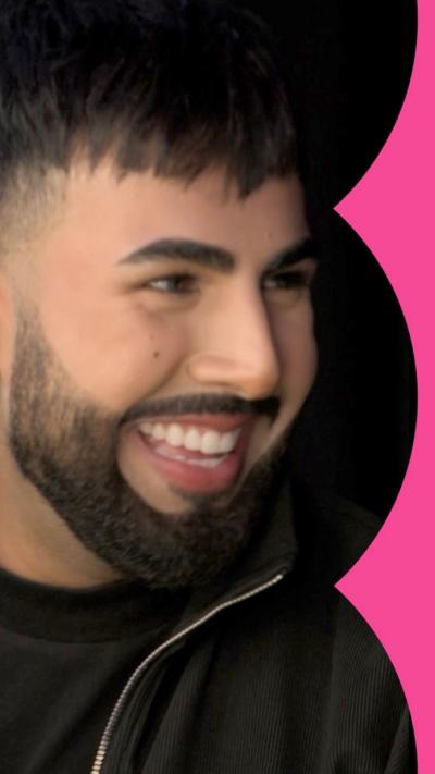 A man, Glow Up contestant, smiling to someone off camera, wearing make-up.