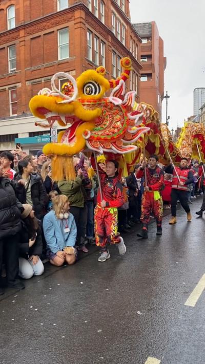 Vibrant dragon costume leading a parade, adorned with bright colours and intricate patterns, surrounded by a cheering crowd.