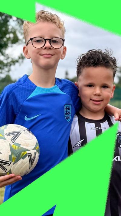 Two children wearing football jerseys smile to the camera. One of them is holding a football.