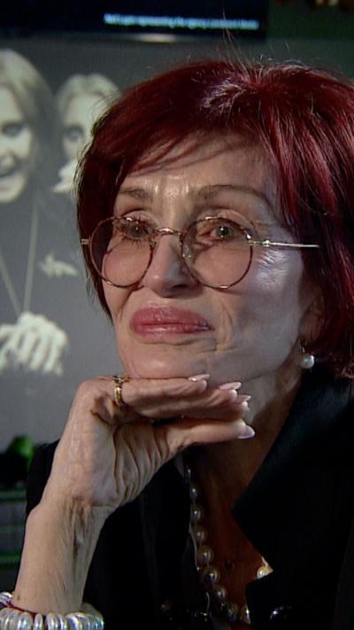 A woman is smiling off camera. Sharon Osbourne has red hair, thin rimmed glasses and red lips. Also wearing a black jacket and pearl necklace.