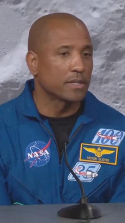 Astronaut in a blue suit with logo patches speaks into a microphone with a Moon background behind him.