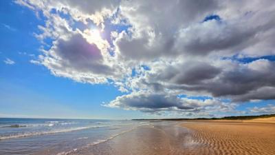 Huge white and grey clouds over golden sand, and shallow sea, with blue sky 
