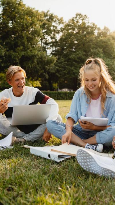 A man with blonde hair in a white jumper and woman with blonde hair in a light blue shirt studying in a park. 