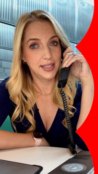 A vertical image of a woman with blonde wavy hair, sitting at a desk and holding a telephone
