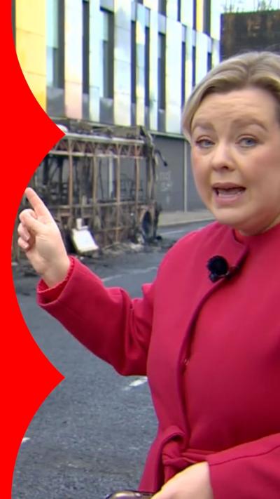 BBC correspondent Sara Girvin wearing pink coat and pointing at burned down bus