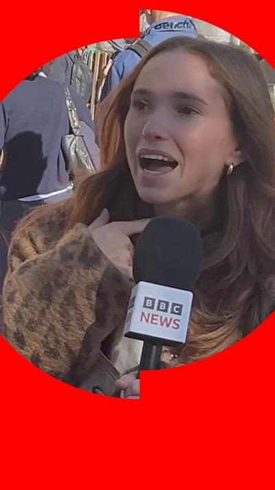 A female student points to her throat while speaking into a BBC News mic. She has long red hair and wears a leopard skin coat 