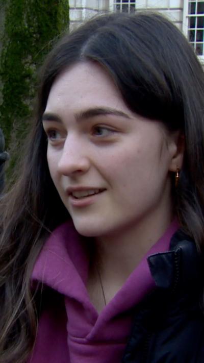 A woman with long brown hair wearing a dark coat over a purple hoody