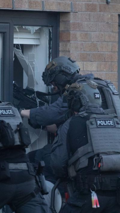Several armed police smashing in through the ground floor window of a brick house.