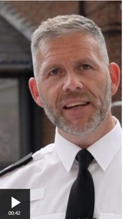 Head and shoulders shot of a man speaking to the camera. He has short greay hair and a grey beard, and is wearing a whte shirt with a black tie. 