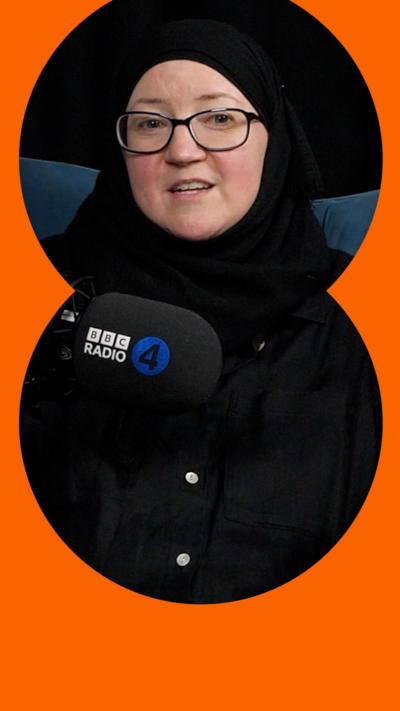 A woman wearing a black hijab and black shirt sits on a teal couch. She is wearing glasses. There is a BBC microphone in front of her. 