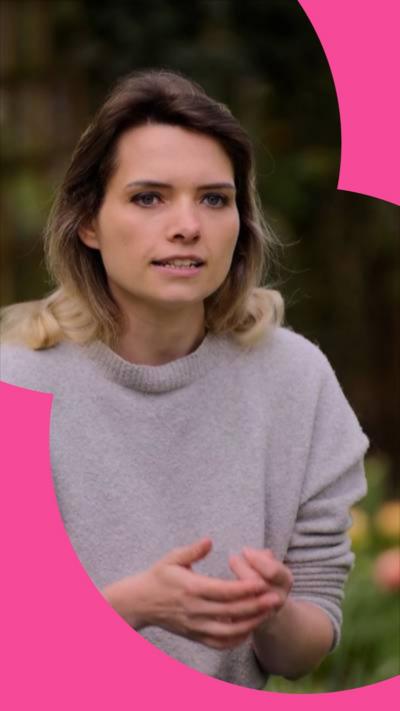 A woman in a lilac jumper sits in a garden. She has long brown hair with blonde highlights