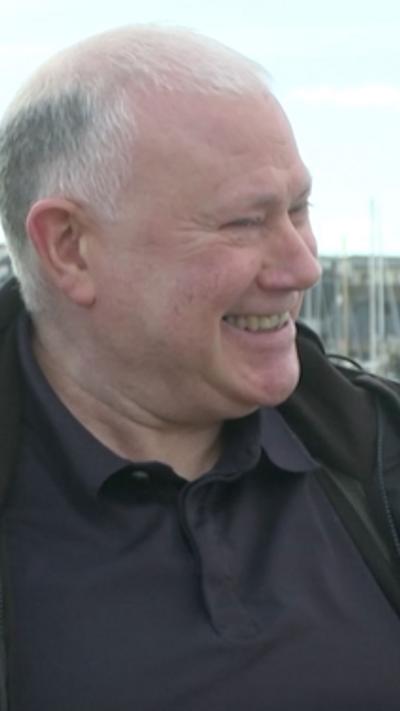Man smiles looking to the right of the camera. It is a head and shoulders shot, he wears a black polo shirt and jacket and has white, balding hair.