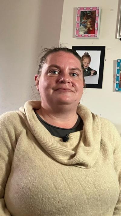 A woman smiles gently while posing for a photo. She is in her home and there are photos on the wall behind her. She is wearing a beige jumper with a rolled neck.