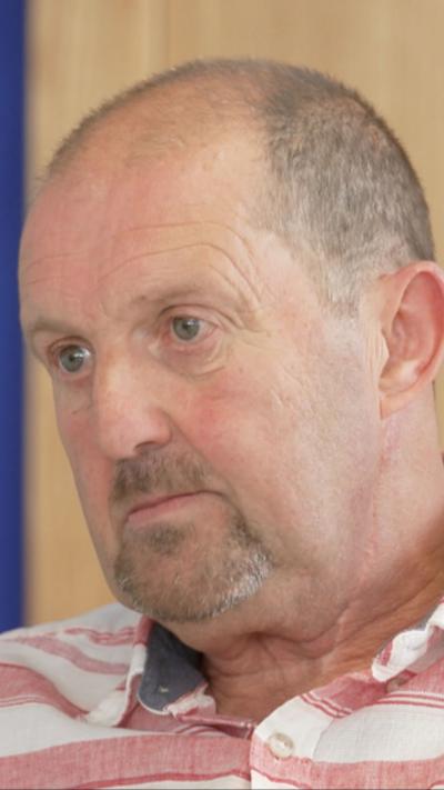 Cliff Donaldson, a man with short, greying hair and a beard, being interviewed for TV in an office setting.  He is wearing a white and pink stripped shirt which is open at the collar.