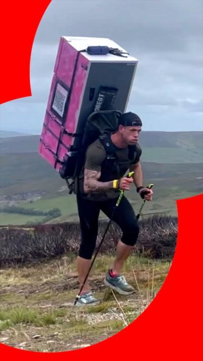 Matt Jones walking on the hills with pink fridge freezer on his back in leggings and khaki green t-shirt