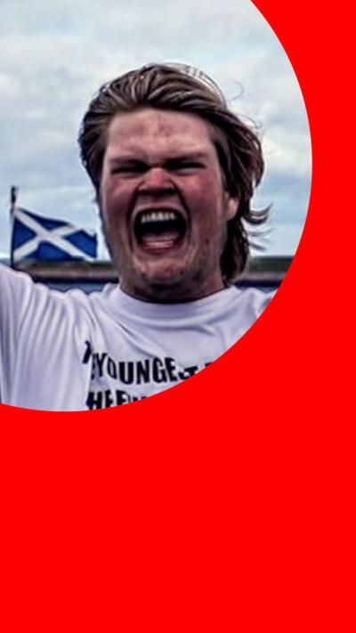 Marcus, aka The Hull Boy, with an expression of joy while holding up a red ribbon, standing by a sign saying 'Land's End 874m'