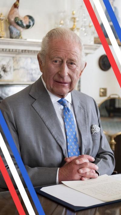 King Charles III, wearing a blue tie and grey suit, looks directly down the camera lens, a radio microphone is hangs about him ready to give his address
