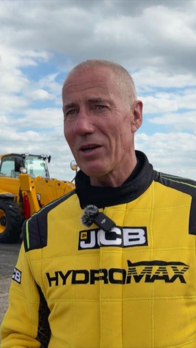 A man with short cropped hair wears a yellow jacket and a clip-on microphone. He is standing in front of a yellow vehicle, with a blue and cloudy sky behind.