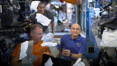 White packages float past smiling astronauts inside the ISS