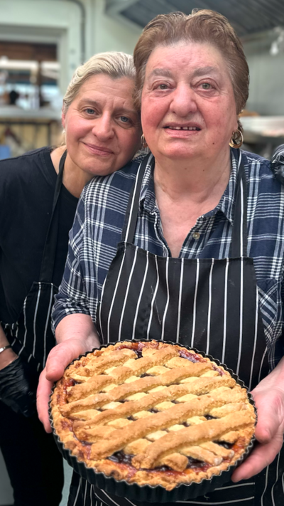 A older and younger woman embrace inside a kitchen while holding a cherry pie. 
