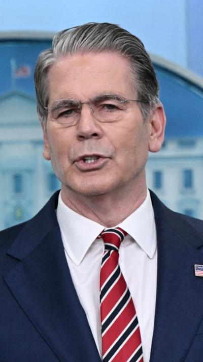 Grey-haired man wearing glasses, a red and blue striped tie and a blue blazer speaks to reporters from the White House briefing room.