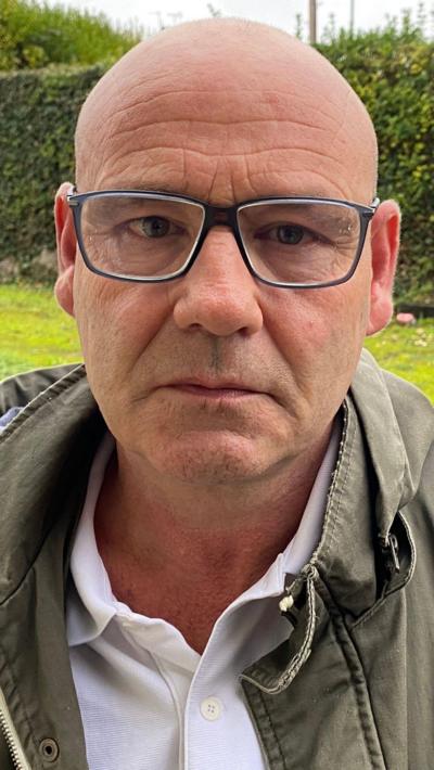 A bald man with black glasses in a green jacket and white shirt