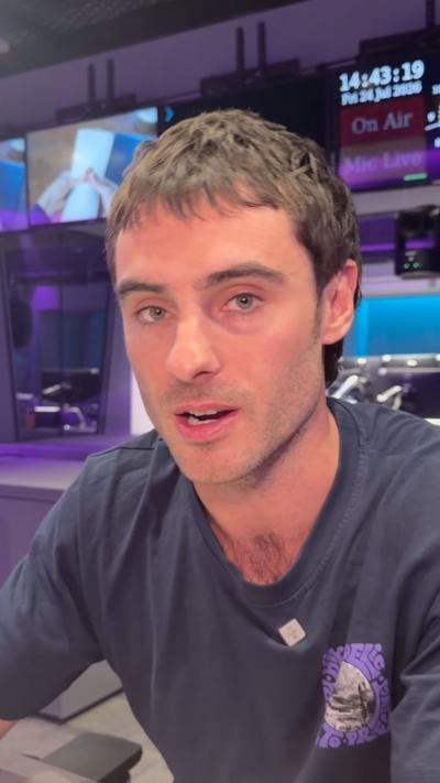 A man with dark hair looks into the camera. He is wearing a dark T-shirt and behind him is a TV screen and a clock.