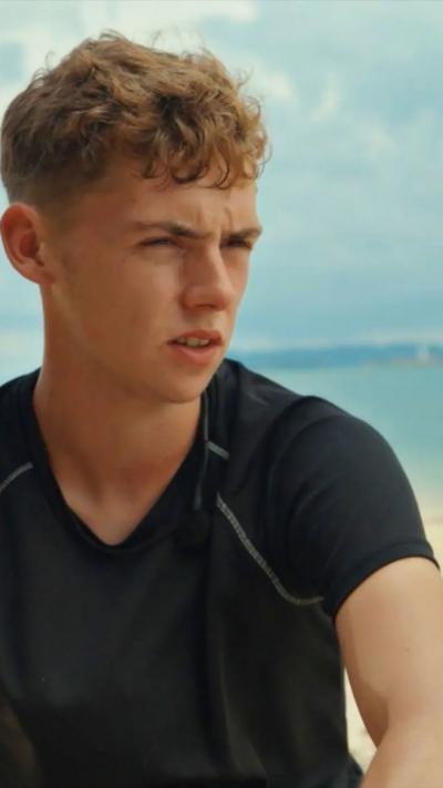 A man with short brown hair wearing a black t-shirt looks away from the camera, with the sea, a beach and a blue sky in the background