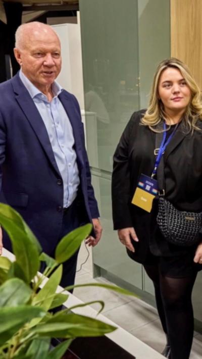 A man in a blue suit and shirt with no tie stands next to a woman wearing a black outfit and a lanyard