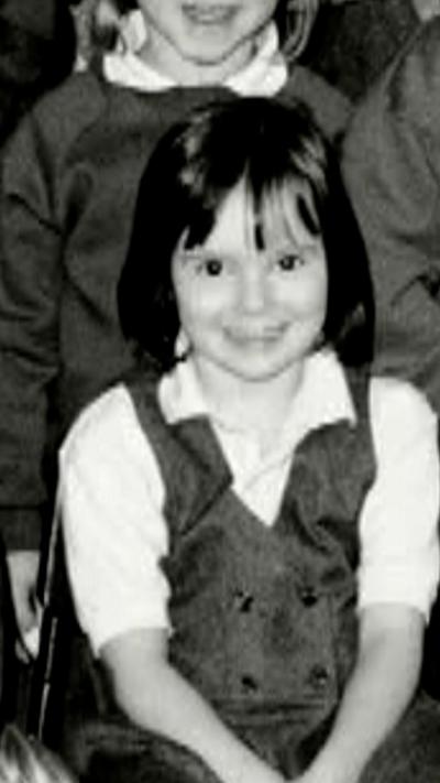A young schoolgirl with short hair and a fringe is smiling at the camera. She is wearing a pinafore dress.