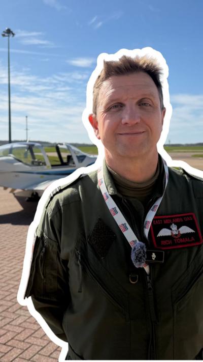 Pilot, wearing a khaki green flight suit, is stood in front of a white aircraft. Blue sky with patchy clouds in the background.