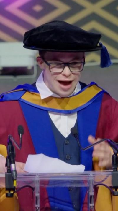 A man stands at a lecture wearing red blue and yellow robes. He wears black rimmed glasses and has a mortar board on his head.