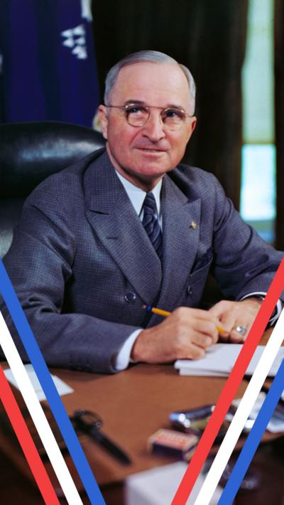A colour photo of US President Harry Truman wearing a grey suit, tie and glasses and smiling towards the camera. He is sat on a black leather chair behind a wooden desk while holding a yellow pencil above a notepad, as if he is about to write something on the paper. There are windows with curtains and a blue flag draped behind him.