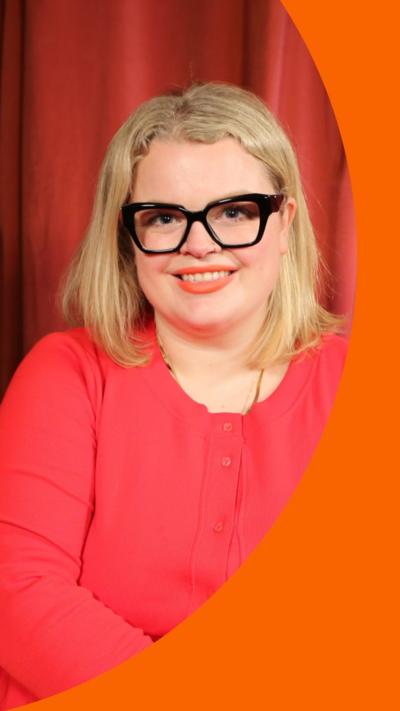 A woman with shoulder-length blonde hair looks into the camera. She is wearing a bright pink top and glasses with black frames.