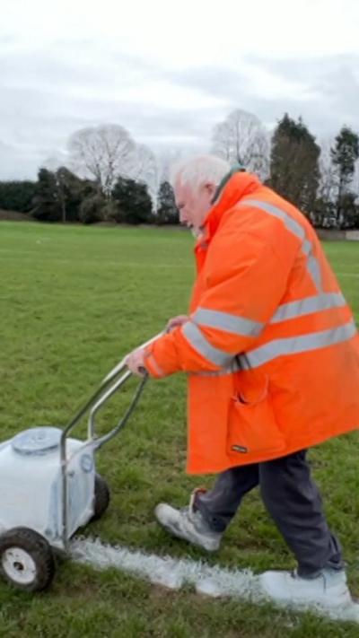 A man in an orange jacket painting white lines on a sports pitch. He has grey hair and is pushing a white machine from right to left