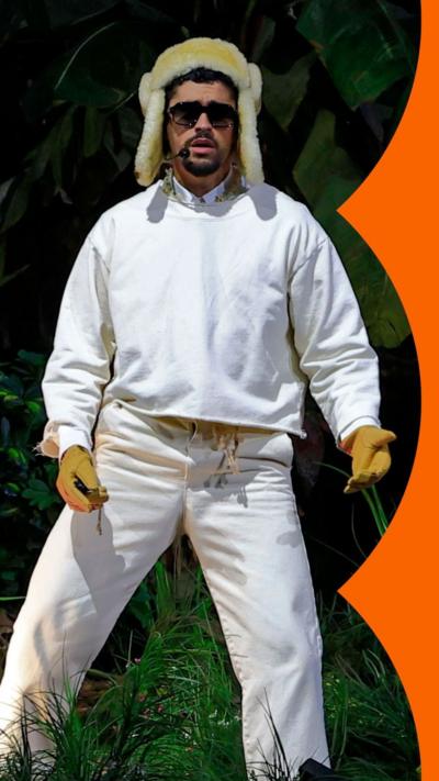 Bad Bunny performs on stage. He wears sunglasses, a woolly hat with ear flaps, and white top and trousers