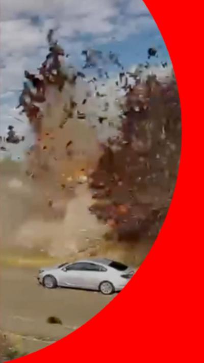 Debris flying through the air mid-explosion, on a tree-lined street with parked cars.