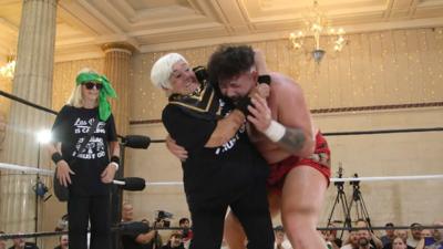 Two women wearing black t-shirts are in a wrestling ring. One of the women is holding a male wrestler with red shorts in a headlock. In the background there are columns, chandeliers and fairy lights covering the cream walls. There are also cameras and members of the audience watching on.  